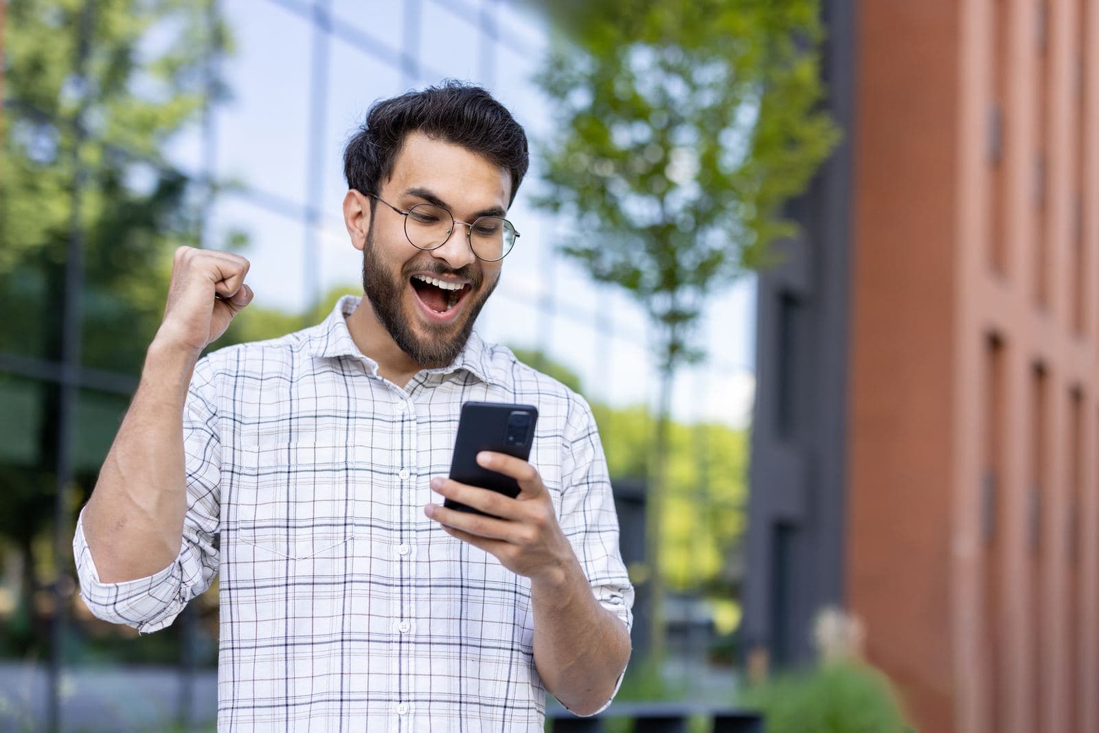 A young man smiling and raising his fist as he reads good news on his phone
